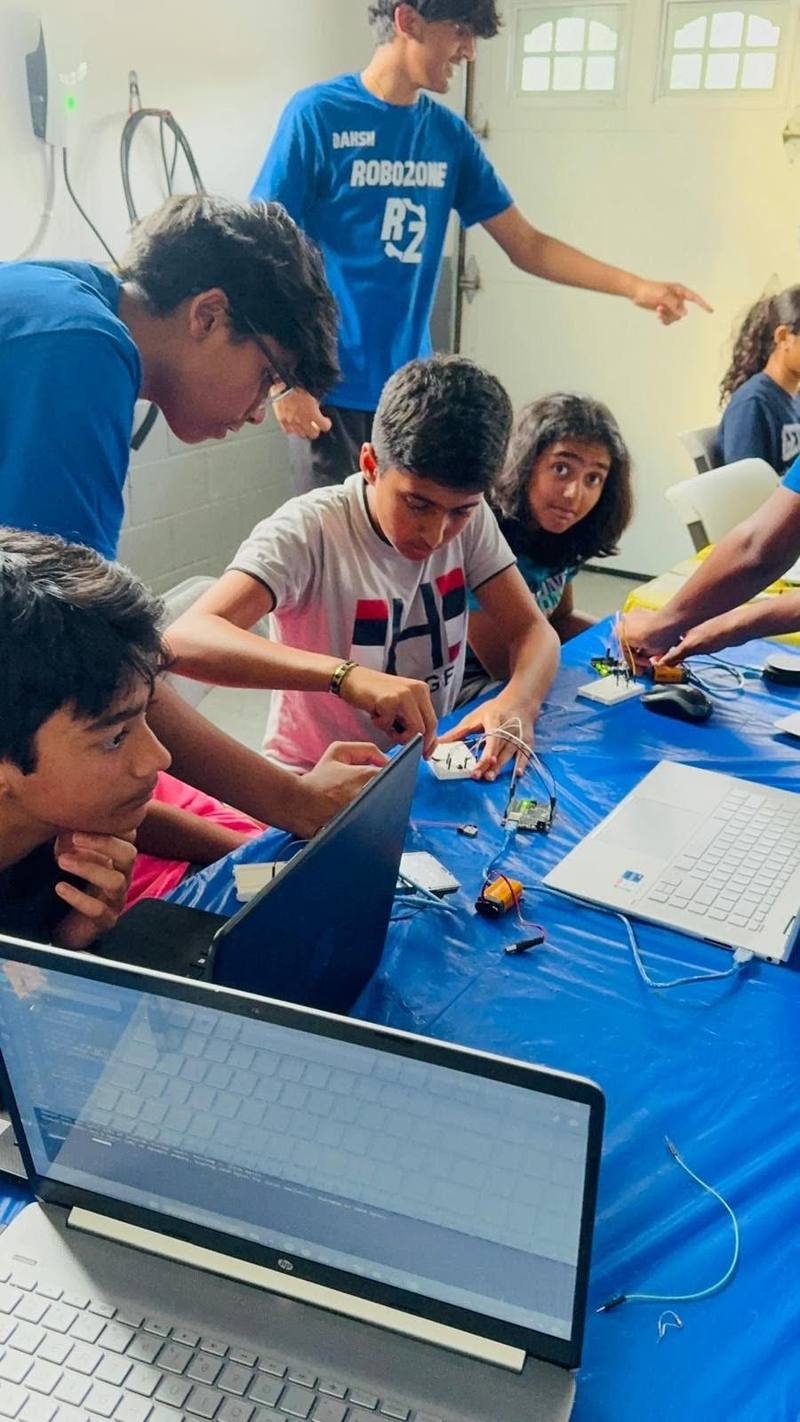 A crowded workshop table: students at laptops and breadboards while RoboZone mentors lean in and point.