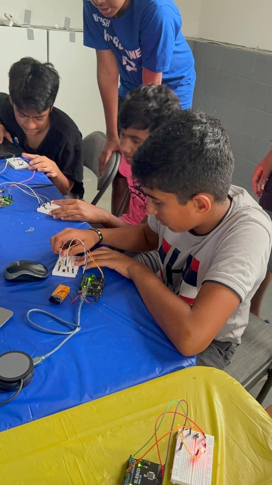 A student pushes wires into a breadboard connected to an Arduino Uno, with other students working at the same table and a mentor standing behind them.