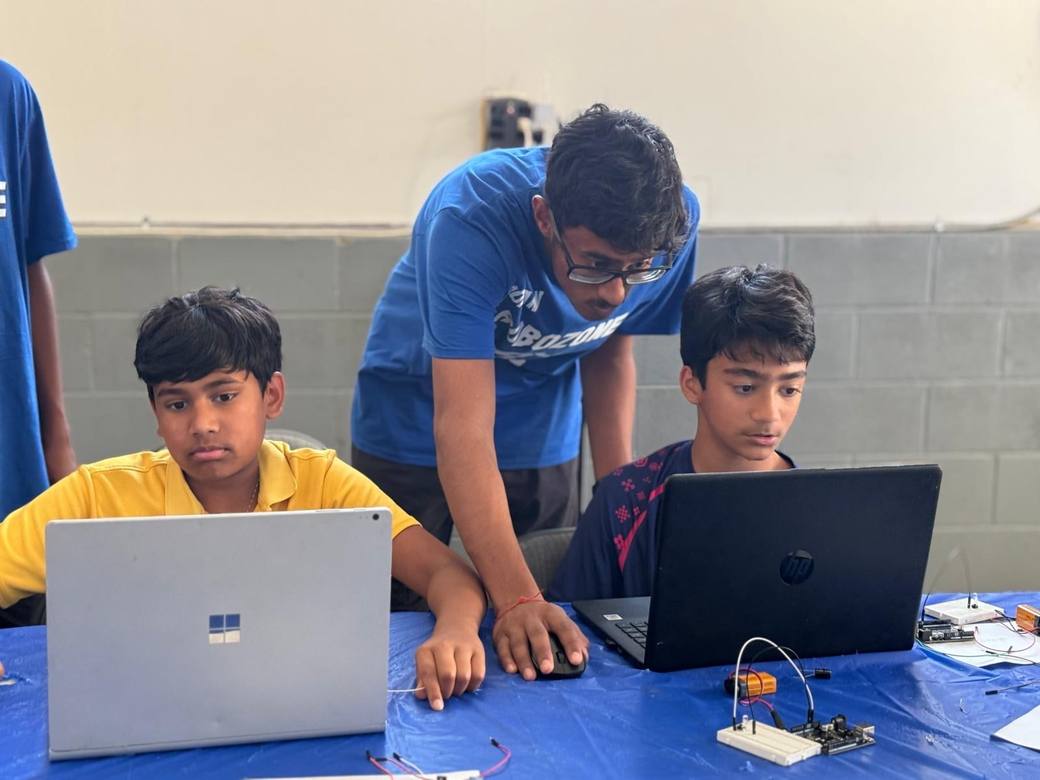A mentor in glasses leans between two students at laptops, with breadboard circuits on the table in front of them.