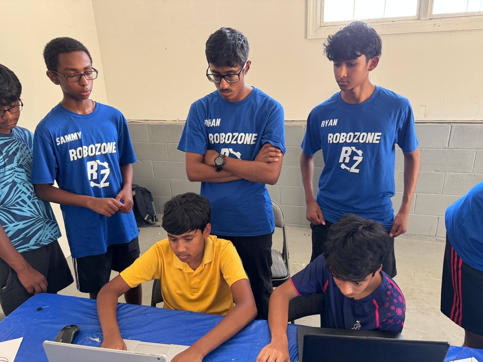 Four mentors in RoboZone shirts stand behind two students working at a laptop.