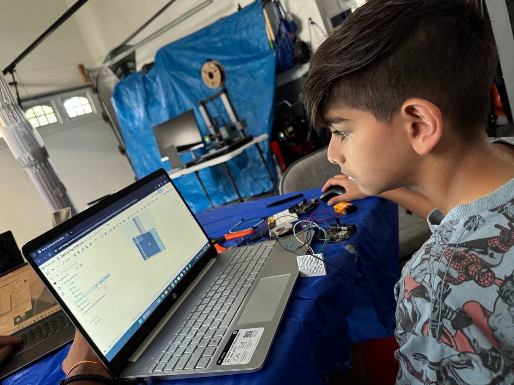 A student looks at a laptop showing a 3D model in Onshape. Arduino boards, a breadboard, and jumper wires sit on the table, with a 3D printer in the background.