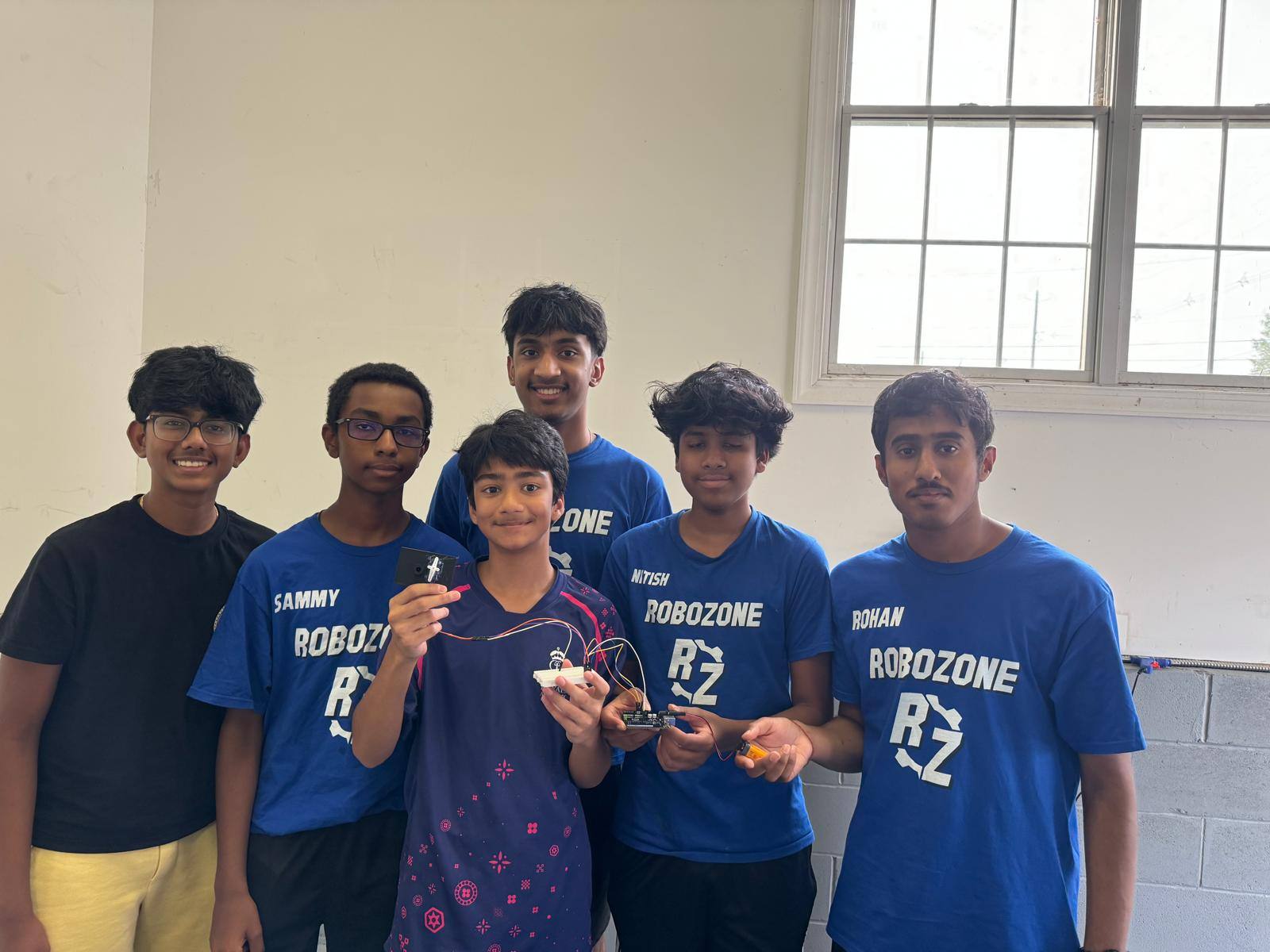 A smiling student holds up a wired project built on a small breadboard. Mentors in blue RoboZone shirts stand around them, holding other parts of the build.