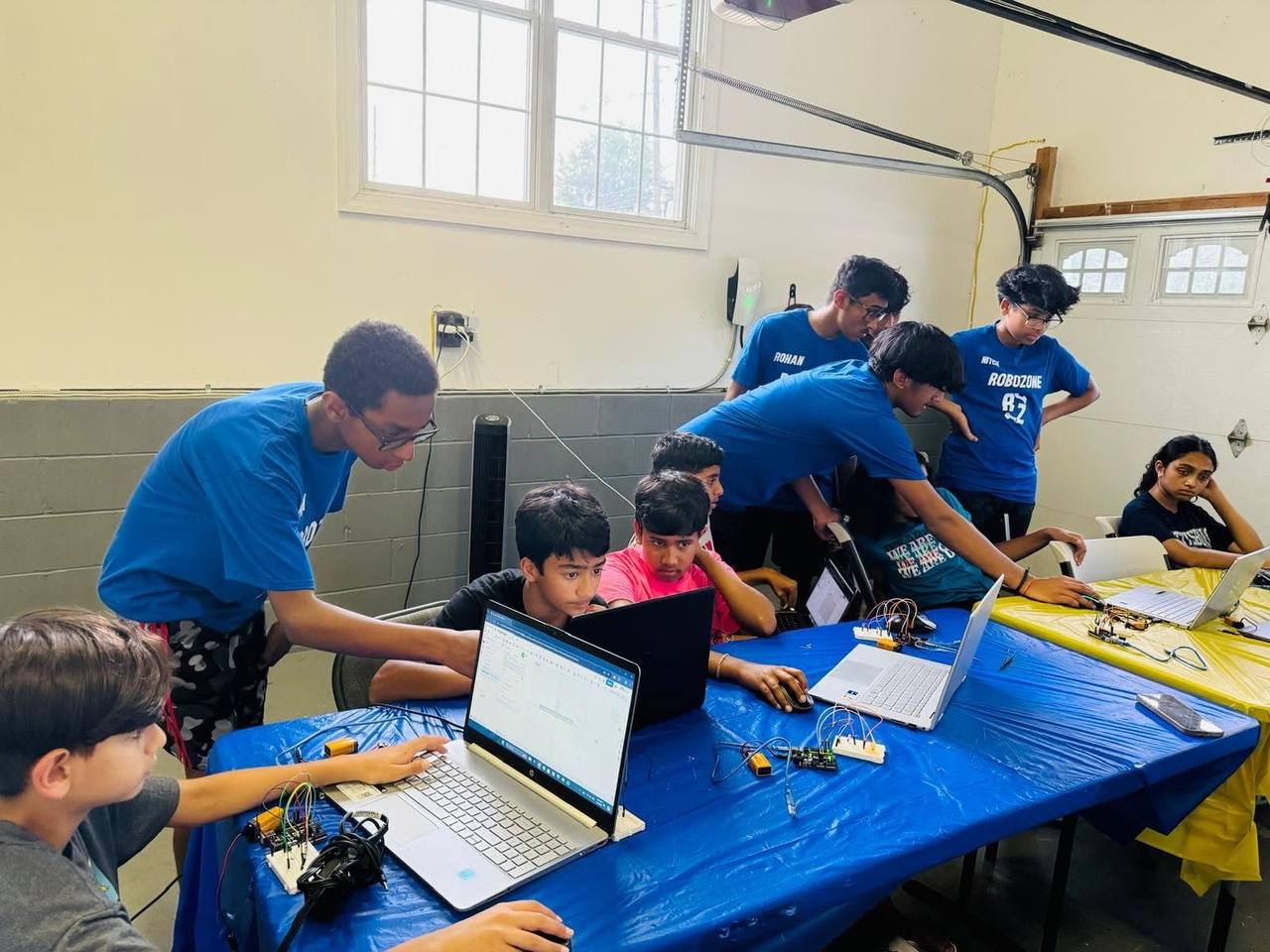 A workshop in progress: mentors in blue RoboZone shirts help students at tables covered with laptops, breadboards, and wires.