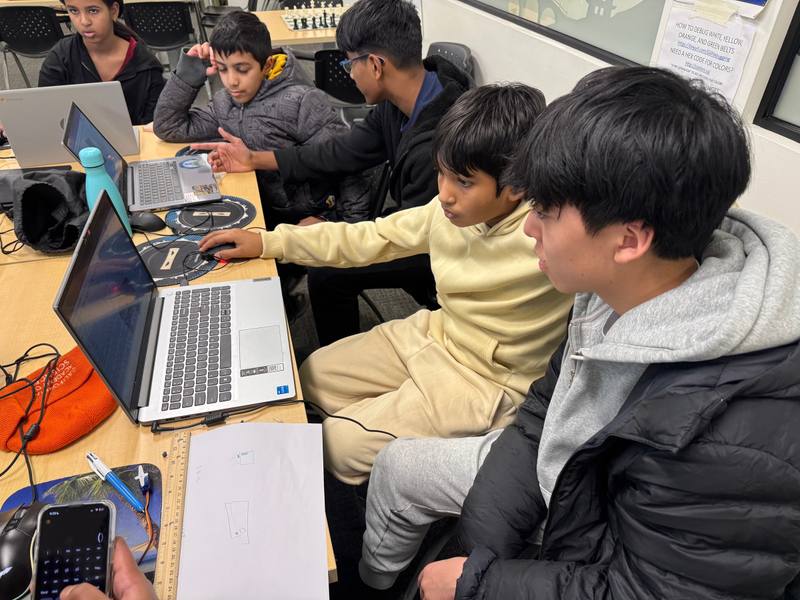 Students working at laptops in a classroom, with a paper sketch and a ruler on the table beside them.