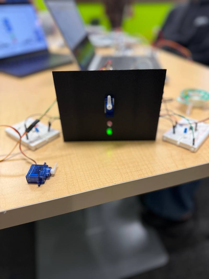 A black 3D-printed panel with a small servo and a lit green LED on the front, beside breadboards and a spare servo.