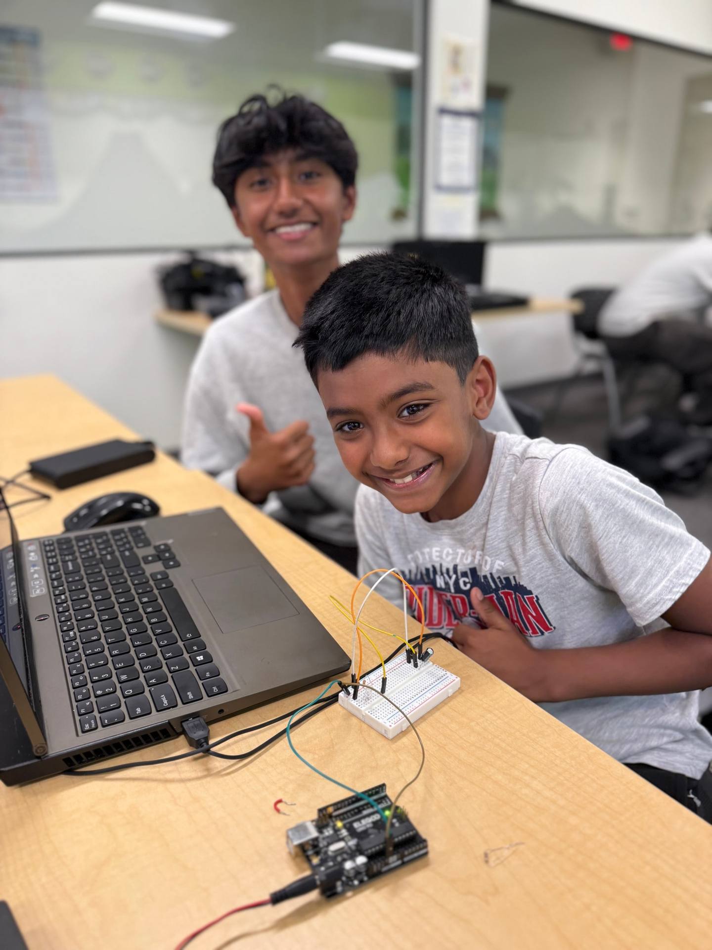 Two smiling students at a workshop table. One gives a thumbs-up behind a breadboard circuit wired to an Arduino Uno.