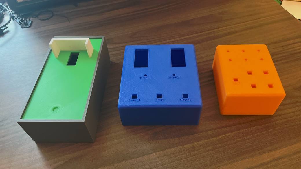 Three 3D-printed cases on a wooden table: a gray one with a green insert, a blue one with cutouts and labels, and an orange one with rows of square holes.
