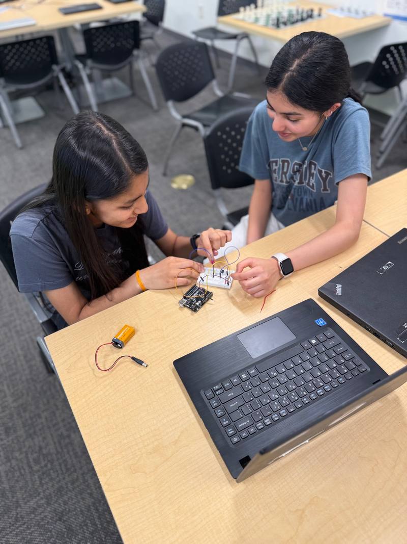 Two students lean in together to place wires on a breadboard connected to a small microcontroller board, with a laptop and a 9V battery on the table.