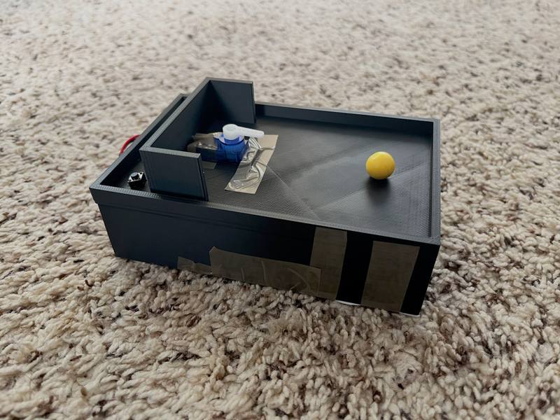 A gray 3D-printed box with a servo mounted in one corner and a small yellow ball inside.